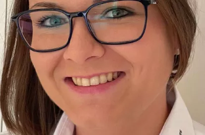 Das Bild zeigt Hattie Huber. Sie ist Ansprechpartnerin für das Jugendrotkreuz im Bezirksverband Oberbayern. Sie hat braune Haare und trägt eine große schwarz umrandete Brille. Auf dem Bild lächelt sie mit geöffnetem Mund.