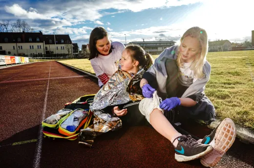 Zwei Juniorhelferinnen, leisten auf dem Schulsportplatz Erste Hilfe bei einer Mitschülerin, die sich am Knie verletzt hat.