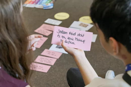 Zwei Kinder sitzen auf dem Boden, Man sieht sie von hinten. Ein Kind hält eine rosafarbene Moderationskarte in der Hand. Darauf steht: "Jedem Kind steht es zu, seine Meinung zu äußern.""
