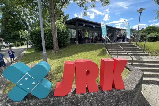 Das Bild zeigt den anmeldebereich des JRK_Landeswettbwewrbs 2024 in berching. im Vordergrund sieht man die großen Styroporbuchstaben des JRK stehen, die aus einem petrolfarbenen kreuz und den roten Lettern "J","R" und"K" bestehen. Im Hinitergund sieht man eine treppe, die zum Eingangsbereich eine Turnhalle führt, an dem sich der Anmeldebereich befindet. Dieser ist durch zwei große petrolfarbenen JRK-Fahnen gekennzeichnet.Einige Teilnehmende stehen dort an.