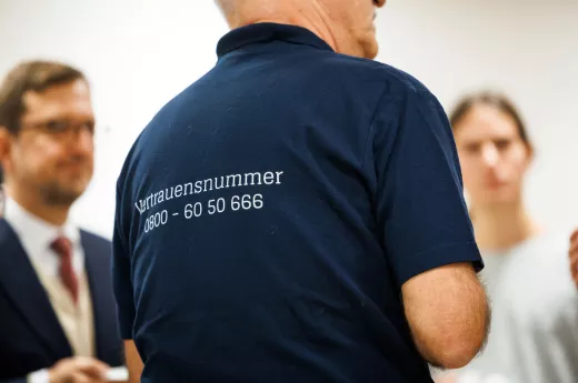 Ein älterer Mann mit grauen Haaren steht im Mittelpunkt des Gesprächs mit mehreren Personen, die im Hintergrund unscharf zu erkennen sind. Auf dem dunkelblauen T-Shirt des Mannes ist die Vertrauensnummer 0800 6050666 sichtbar.