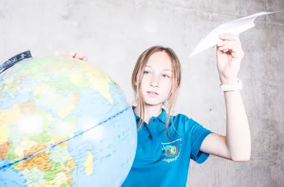 Das Bild ziegte eine Jugendrotkreuzlerin, die einen Papierflioegr in der Hand hält. Mit der anderen Hand dreht sie einen Globus.