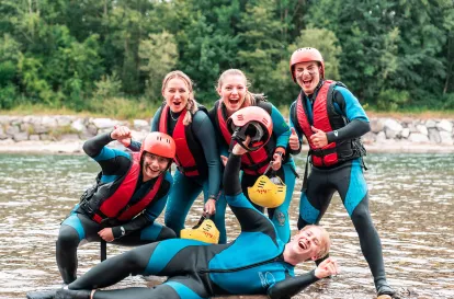 5 Jugendliche, die lachend für ein Foto possieren. Sie stehen in einem seichten Fluss und tragen blau-schwarze Neoprenanzüge und Helme. 