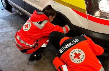 Das BIld zeigt zwei Rettungdienstmitarbeitende, die ein Rad des Rettungswagens überprüfen.