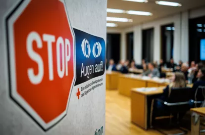Im Vordergrund auf der linken Seite sieht man ein Roll-up mit dem Logo der Initiative 'Stop Augen auf'. Es wird von einem verschwommenen Konferenzsaal im Hintergrund begleitet, in dem Menschen konzentriert einer Podiumsdiskussion folgen.