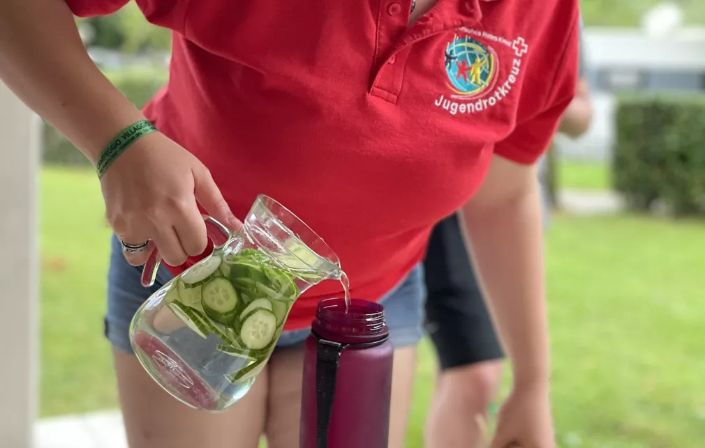 Auf dem Bild sieht man eine Person, die sich aus einer Glaskaraffe Gurkenwasser in eine mitgebrachte Flasche füllt. Die Person trägt ein rotes Polo-Shirt mit Jugendrotkreuz-Logo auf der rechten Brust.